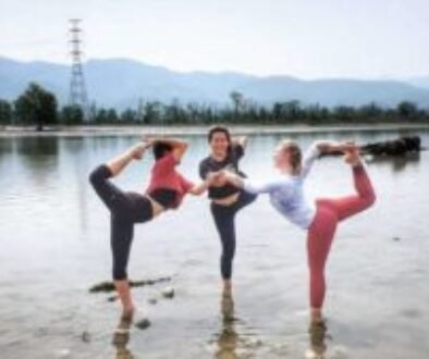 Three yoga teacher training students gracefully perform a yoga pose together with their feet submerged in the sacred waters of the river Ganges.
