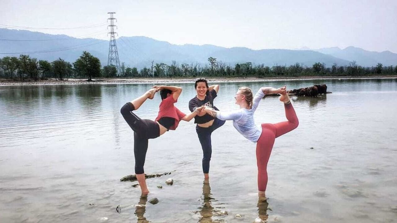 Three yoga teacher training students gracefully perform a yoga pose together with their feet submerged in the sacred waters of the river Ganges.